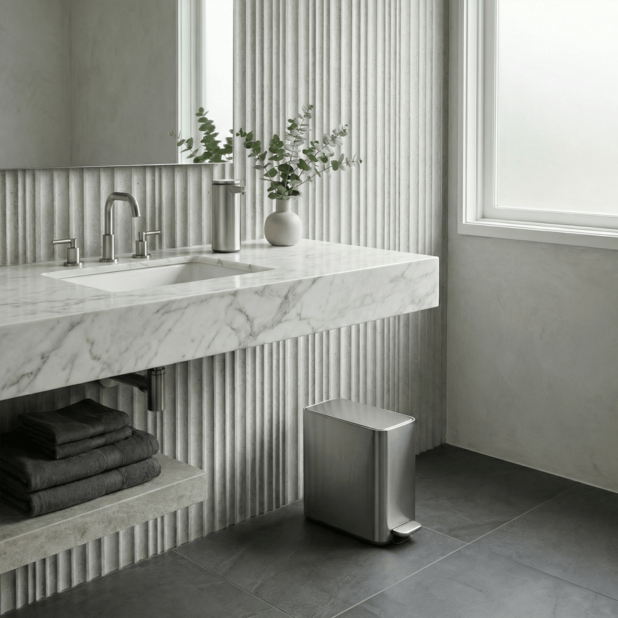Simplehuman's 9L Slim Step Can in Brushed in a modern, simple bathroom setting. Textured walls with stone flooring and a Simplehuman Sensor Pump in Brushed on the marble sink countertop.